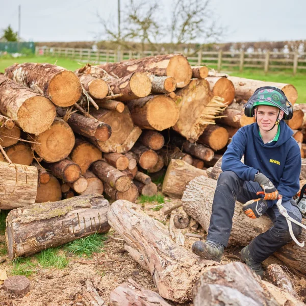 A smiling child in safety gear sitting on logs with a chainsaw.