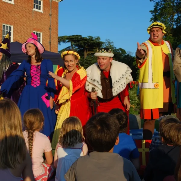 Actors in colourful costumes performing outdoors for an audience of children, in front of a brick building.