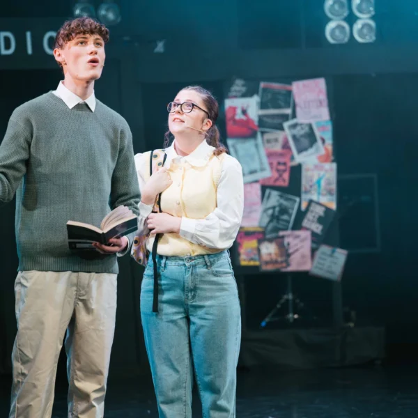 A man and woman on a stage, man holding book and gesturing, woman smiling and looking up.