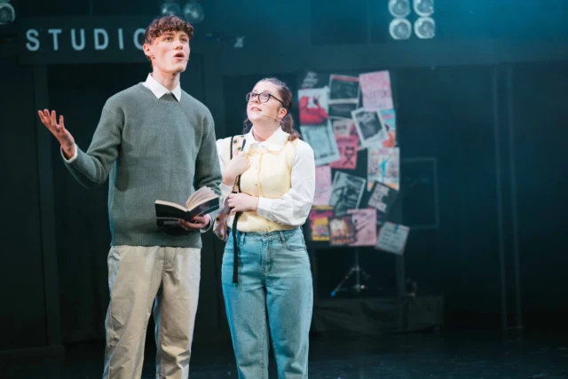 A man and woman on a stage, man holding book and gesturing, woman smiling and looking up.