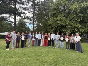 Group of people standing in a line on grass holding certificates, smiling and looking at the camera.