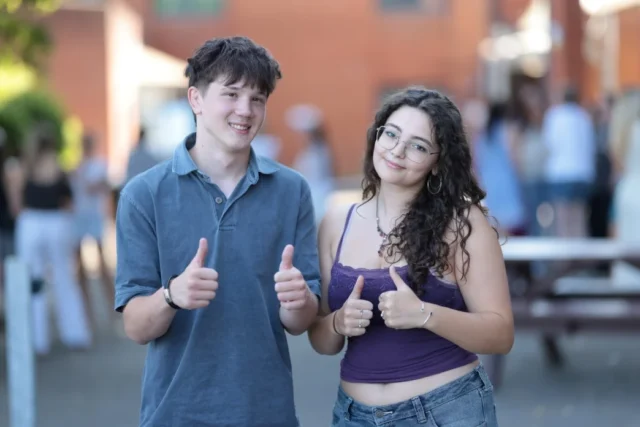 Smiling teenage boy and girl giving thumbs up outdoors near brick school building