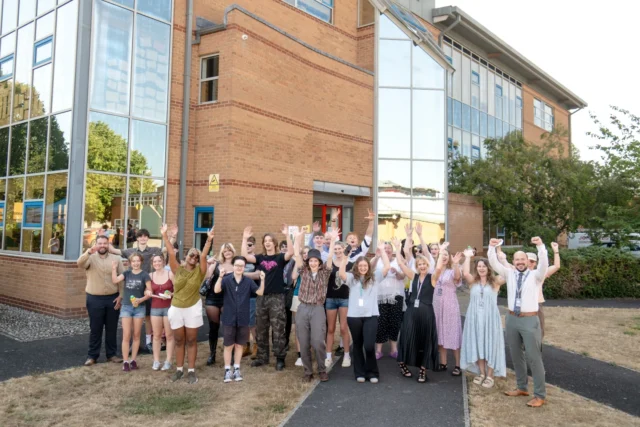 GROUP OF STUDENTS CHEERING OUTSIDE A BRICK BUILDING WITH GLASS WINDOWS.