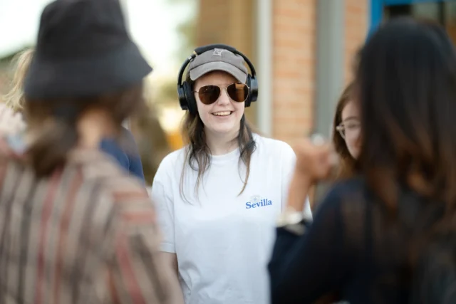 A cheerful woman wearing a cap, sunglasses, and headphones smiles at the camera while surrounded by friends in an outdoor setting.