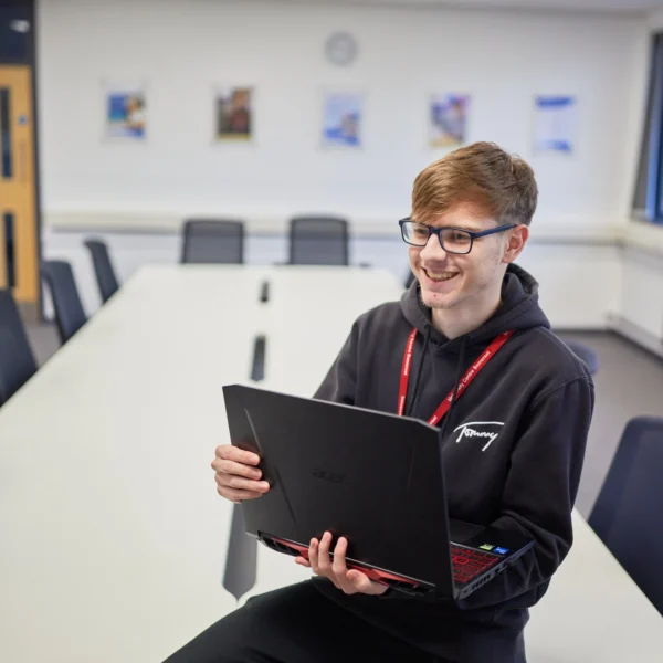Young man sitting on a chair holding a laptop in a classroom or meeting room setting.
