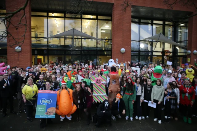 A large group of people gathered outside a building for a fun event, some wearing costumes and holding signs.