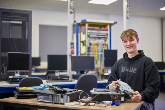 A young man in a black hoodie working on computer hardware in a classroom setting.