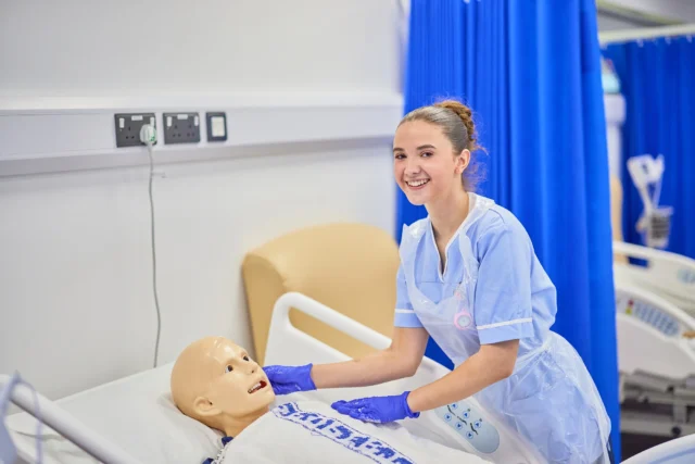A woman in blue scrubs interacts with a mannequin on a hospital bed.