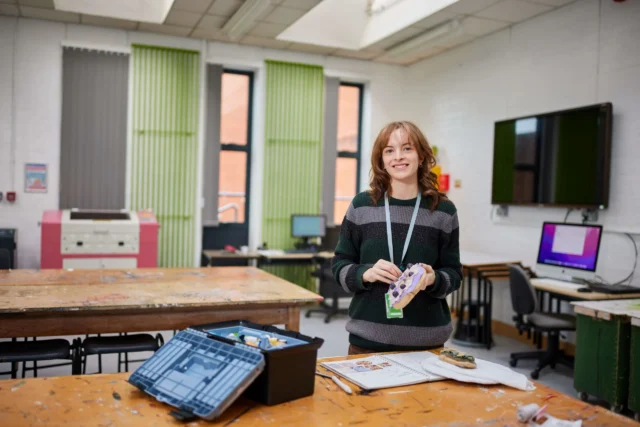 A smiling woman standing in a classroom with a lanyard around her neck.