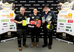 Three men holding electrical equipment in a promotional setting with 'SPARKS SKILLS' and 'ELECTRIC CENTER' banners.