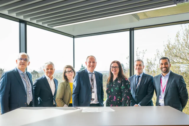 A group of seven business professionals standing around a table in a modern office with large windows.