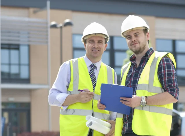 Two construction workers in hard hats and reflective vests looking at blueprints on a clipboard.