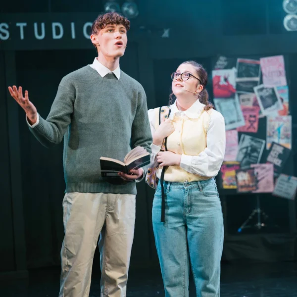 A man and woman on a stage, man holding book and gesturing, woman smiling and looking up.