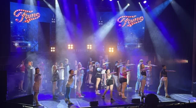 A vibrant stage performance with a group of energetic dancers wearing casual clothing, illuminated by bright lights and fog effects.