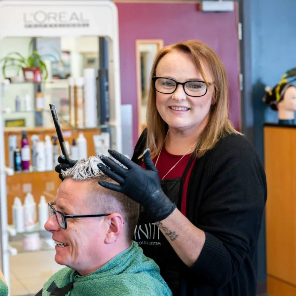 A professional hairdresser applying highlights to a client's hair in a vibrant salon setting with L'Oréal branding visible in the background.