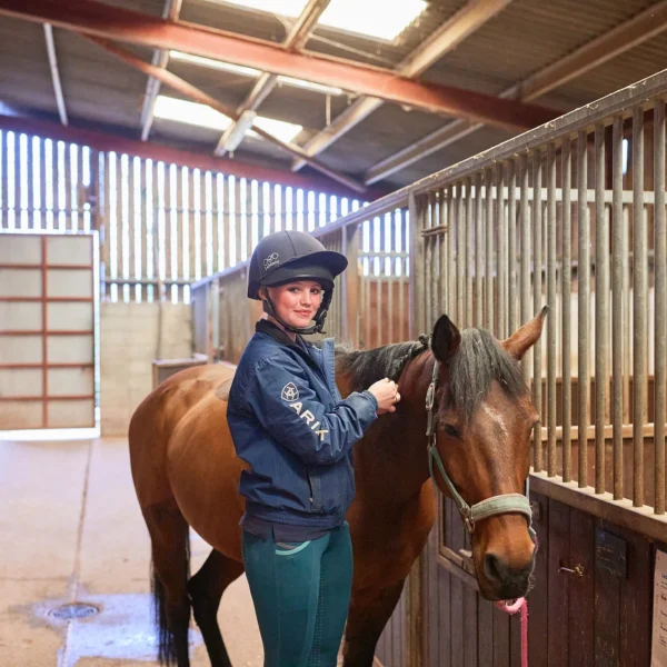 Woman in riding gear standing with a brown horse in a stable.