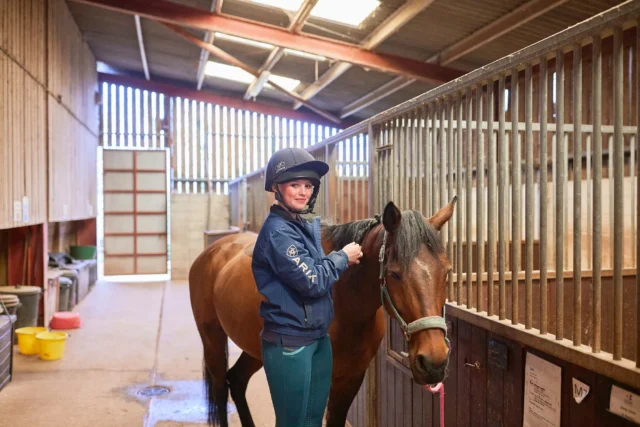 Woman in riding gear standing with a brown horse in a stable.