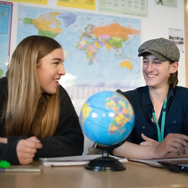 Two young women sitting at a desk with a globe, looking at each other and smiling.