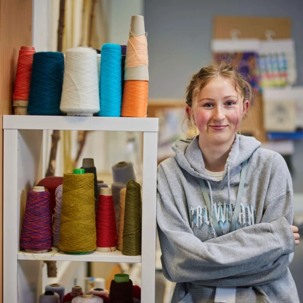 Young girl with arms crossed standing next to a shelf of colorful threads.