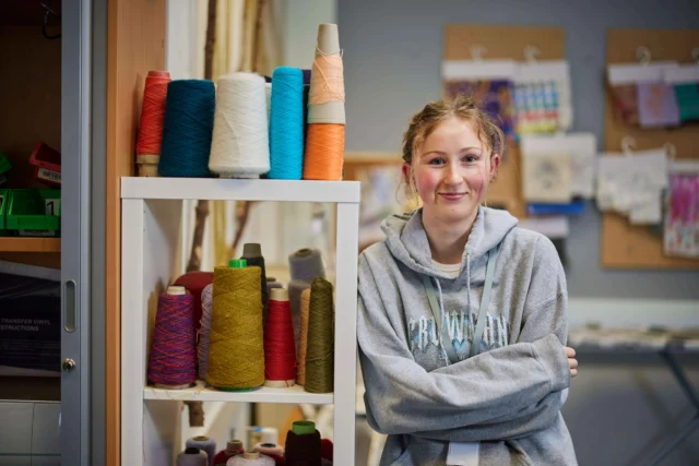Young girl with arms crossed standing next to a shelf of colorful threads.