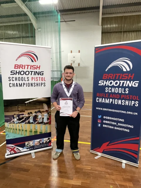 Man holding certificate standing between two British Shooting Schools Pistol Championships banners.