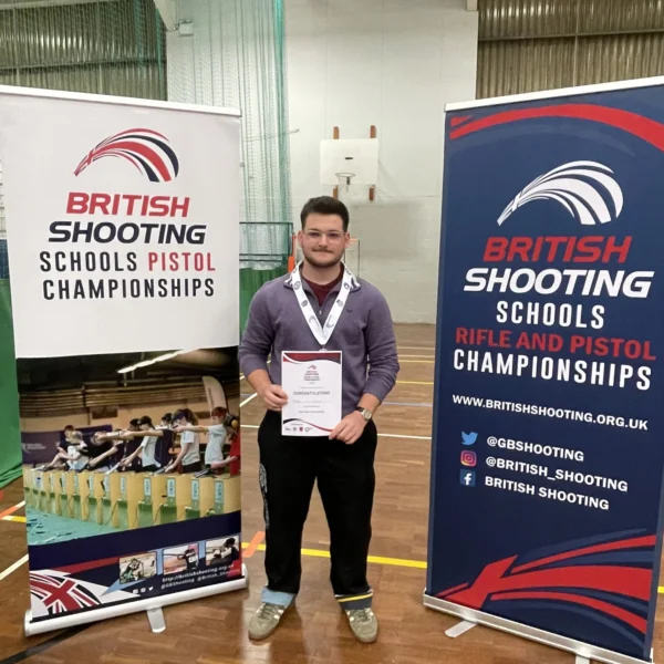 Man holding certificate standing between two British Shooting Schools Pistol Championships banners.