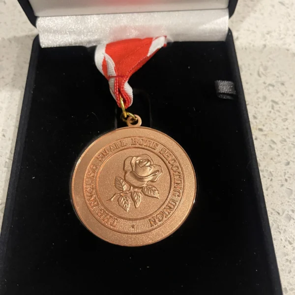 A bronze medal with a rose emblem on a red and white ribbon in a black velvet box.