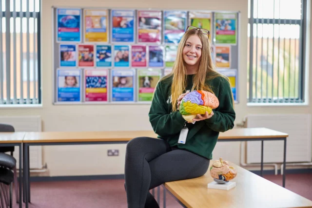 A young woman holds a model of a human head in a classroom.