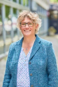 A woman with short blonde hair and glasses smiling outside, wearing a blue blazer and a white blouse with blue dots.