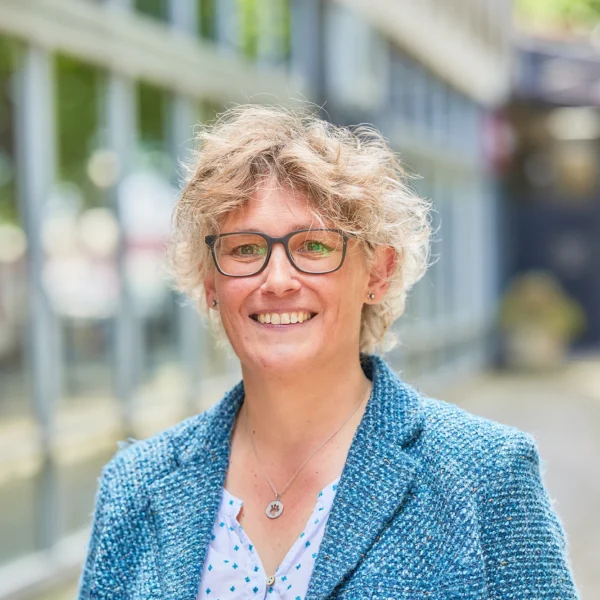 A woman with short blonde hair and glasses smiling outside, wearing a blue blazer and a white blouse with blue dots.