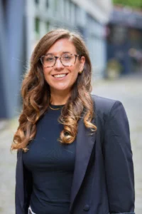 A smiling businesswoman with long brown hair and glasses wearing a navy blue blazer.