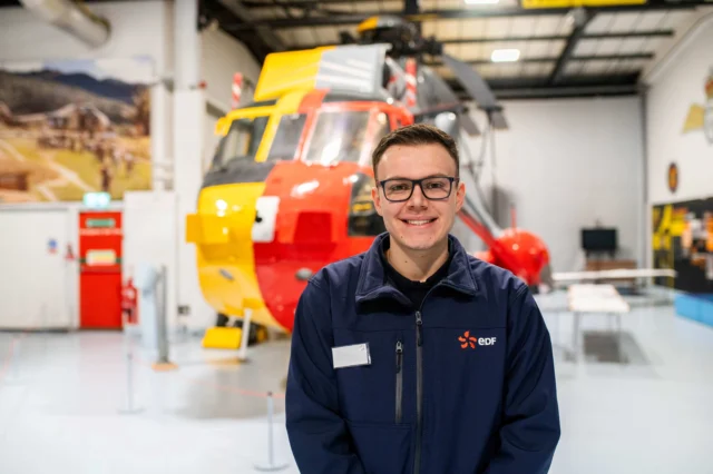 A man in glasses and a blue jacket standing in front of a helicopter.