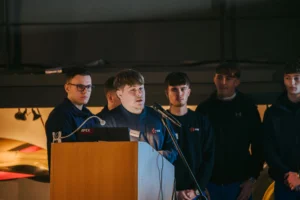 Young man speaking at a podium with four others standing behind him.
