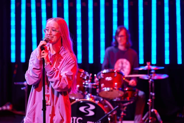 Woman singing into a microphone on stage, with a drummer in the background. The singer has long blonde hair and is wearing a pink jacket.