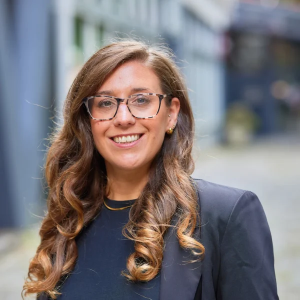 A smiling woman with long brown hair and glasses wearing a black top and blazer.