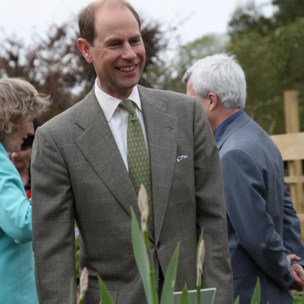 Prince Edward in a green suit and tie stands in a garden with couple.
