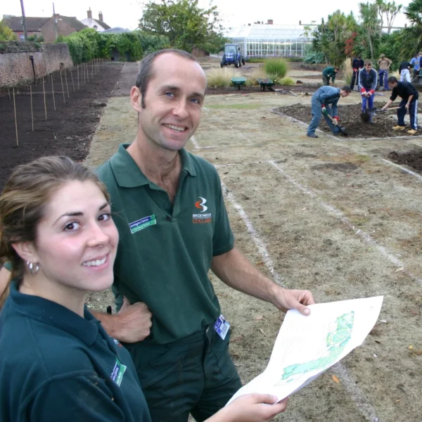 A man and woman in green uniforms holding a garden plan, with people working in the background.