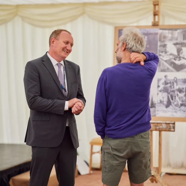 Two men in a room with a framed picture on an easel behind them, one in a suit and the other in casual clothing.
