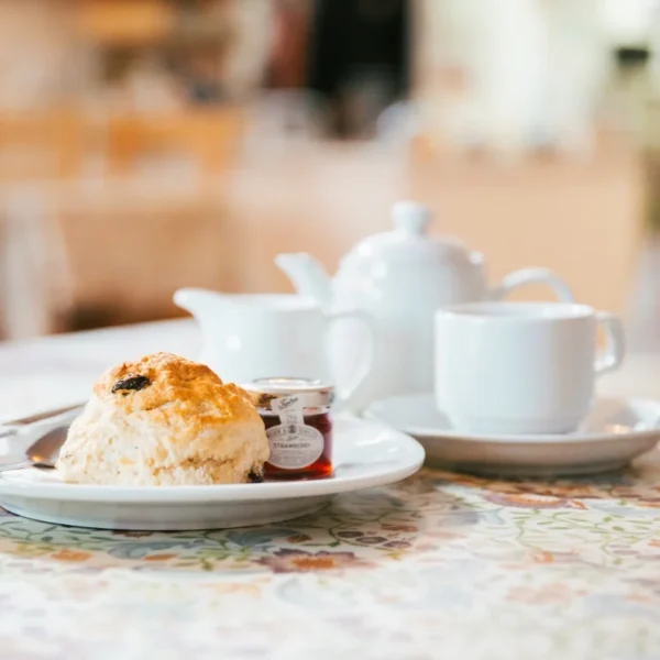 A scone on a plate with a cup and teapot in the background on a table.