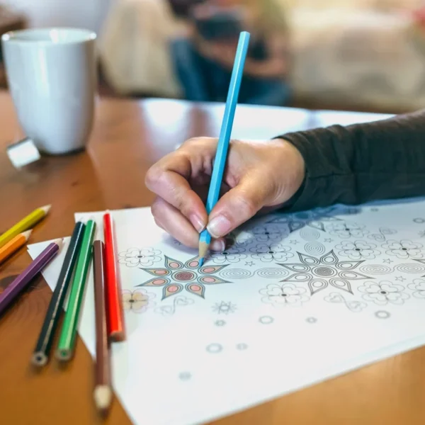 A hand coloring a mandala pattern with colored pencils on a wooden table.