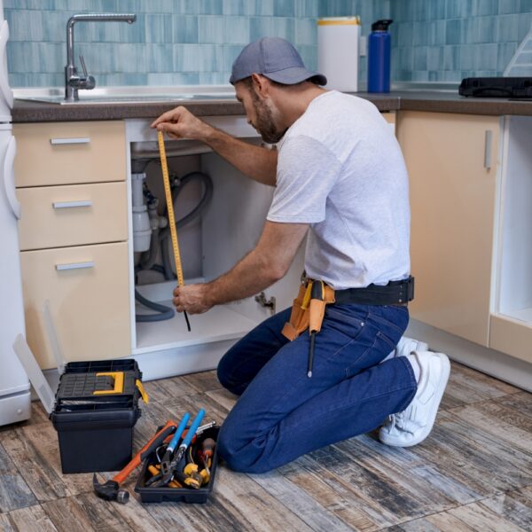 A man in a white shirt and blue jeans kneels on a wooden floor in a kitchen, using a tape measure to work under a cabinet. A toolbox and tools are nearby.