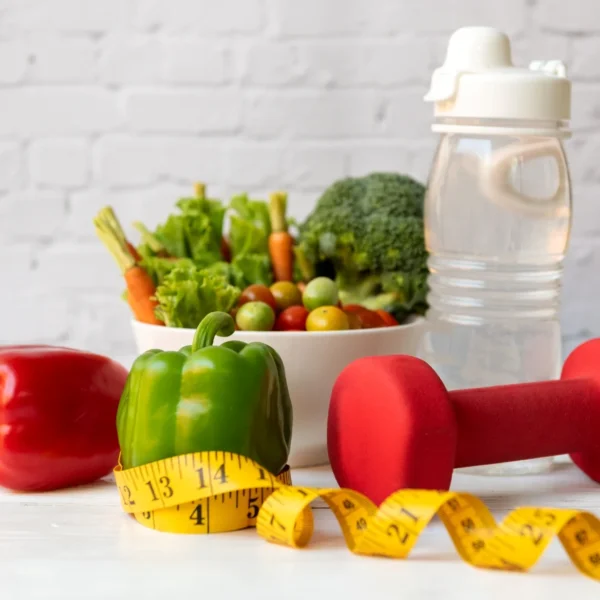 Red dumbbell, yellow measuring tape, green bell pepper on a white surface with a bowl of fresh vegetables and water bottle in the background.