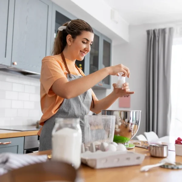 A woman in an apron baking in a bright kitchen, pouring ingredients into a stand mixer surrounded by flour, eggs, and berries.