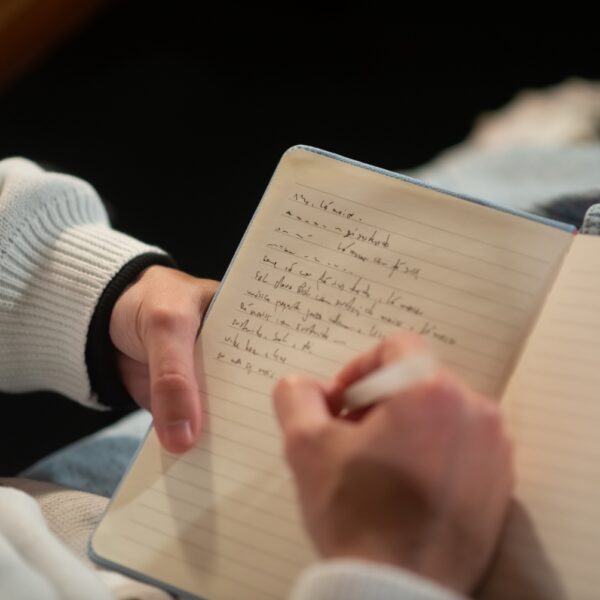 A person writing in a notebook, focusing on their hands and the open notebook on their lap, wearing a white sweater and jeans.