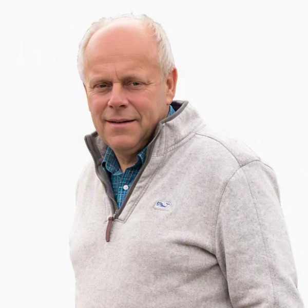 Portrait of a smiling senior Caucasian man wearing a grey fleece sweater with a brown zipper against a white background.