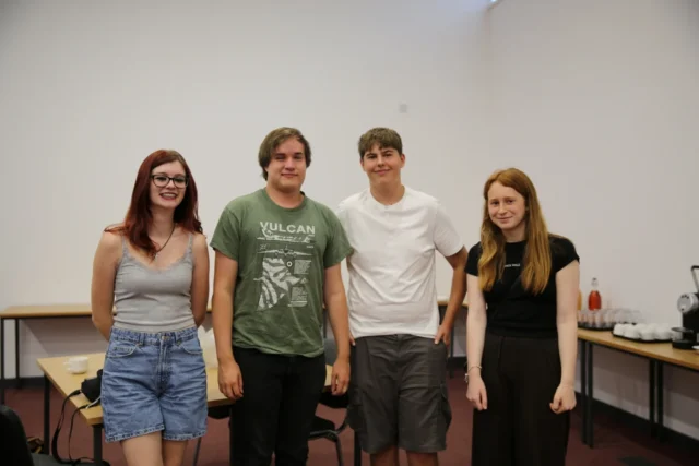 Four individuals posing together in a room, with tables visible in the background.