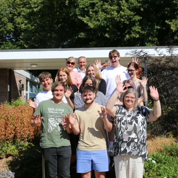A group of people smiling and waving in front of a building with lush greenery surrounding them.