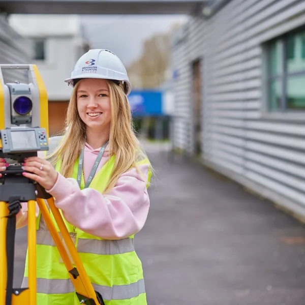 Young surveyor in high-visibility vest operating a theodolite on a tripod outside a modern building.