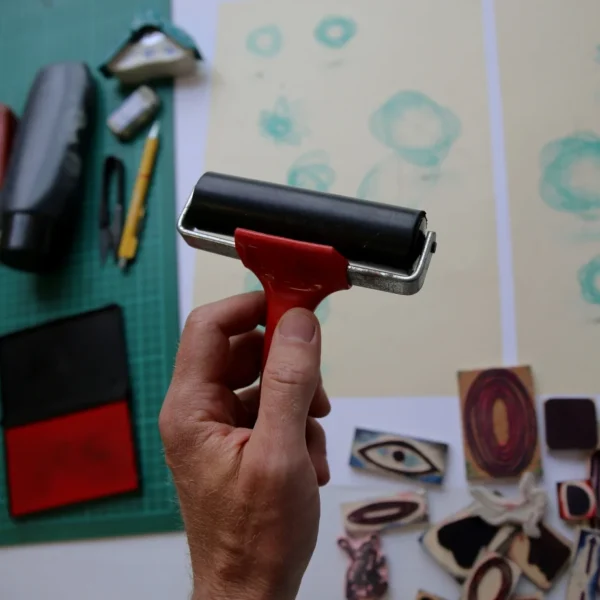 Hand holding a roller brayer over a green cutting mat with ink pads and rubber stamps nearby.