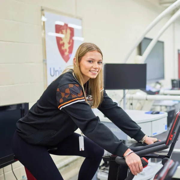 A female sport student on an exercise bike in the UCS Sports Performance Lab. She is wearing black sportswear and is smiling at the camera.